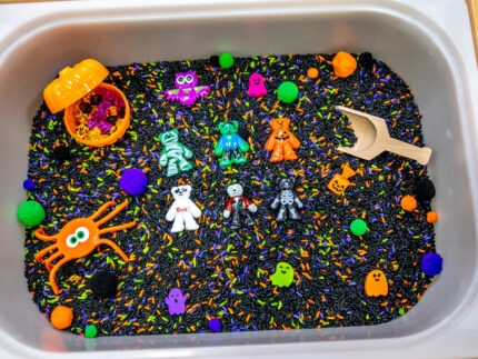Halloween Bear Sensory Bin with colorful Halloween rice, costumed bears, ghosts, spider, pom-poms, gems, pumpkin container and wooden scoop