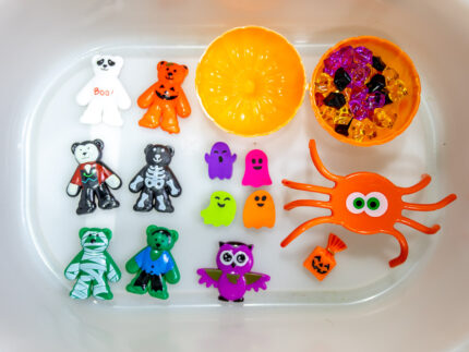 Halloween Bear Sensory Bin with colorful Halloween rice, costumed bears, ghosts, spider, pom-poms, gems, pumpkin container and wooden scoop