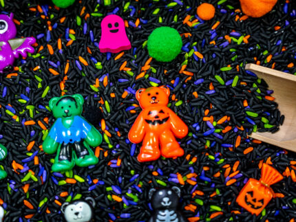 Halloween Bear Sensory Bin with colorful Halloween rice, costumed bears, ghosts, spider, pom-poms, gems, pumpkin container and wooden scoop