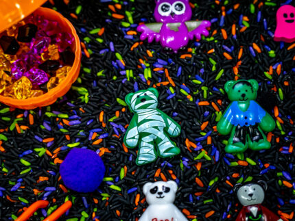 Halloween Bear Sensory Bin with colorful Halloween rice, costumed bears, ghosts, spider, pom-poms, gems, pumpkin container and wooden scoop