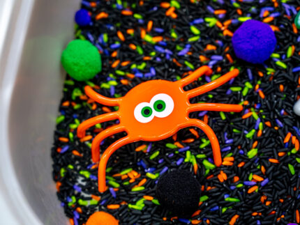 Halloween Bear Sensory Bin with colorful Halloween rice, costumed bears, ghosts, spider, pom-poms, gems, pumpkin container and wooden scoop