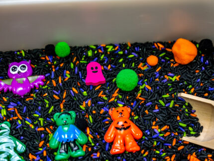 Halloween Bear Sensory Bin with colorful Halloween rice, costumed bears, ghosts, spider, pom-poms, gems, pumpkin container and wooden scoop
