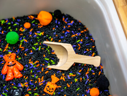 Halloween Bear Sensory Bin with colorful Halloween rice, costumed bears, ghosts, spider, pom-poms, gems, pumpkin container and wooden scoop