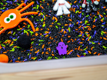 Halloween Bear Sensory Bin with colorful Halloween rice, costumed bears, ghosts, spider, pom-poms, gems, pumpkin container and wooden scoop