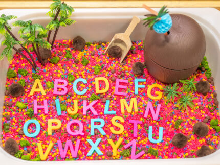 Chicka Chicka Alphabet Sensory Bin with colorful sensory filler, alphabet letters, coconut container, palm trees, pom-pom coconuts and wooden scoop
