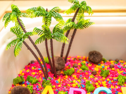 Chicka Chicka Alphabet Sensory Bin with colorful sensory filler, alphabet letters, coconut container, palm trees, pom-pom coconuts and wooden scoop