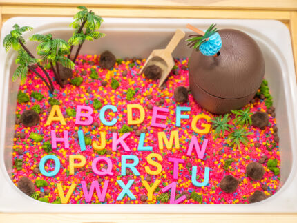 Chicka Chicka Alphabet Sensory Bin with colorful sensory filler, alphabet letters, coconut container, palm trees, pom-pom coconuts and wooden scoop