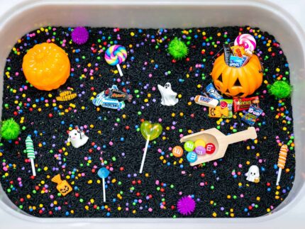 Trick-or-Treat Sensory Bin with black rice, rainbow couscous, pumpkin containers, pretend candy, ghosts, pom-poms, and wooden scoop