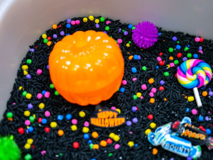 Trick-or-Treat Sensory Bin with black rice, rainbow couscous, pumpkin containers, pretend candy, ghosts, pom-poms, and wooden scoop