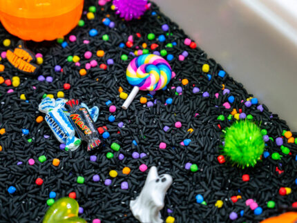 Trick-or-Treat Sensory Bin with black rice, rainbow couscous, pumpkin containers, pretend candy, ghosts, pom-poms, and wooden scoop
