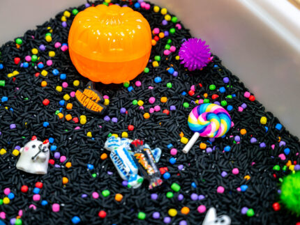 Trick-or-Treat Sensory Bin with black rice, rainbow couscous, pumpkin containers, pretend candy, ghosts, pom-poms, and wooden scoop