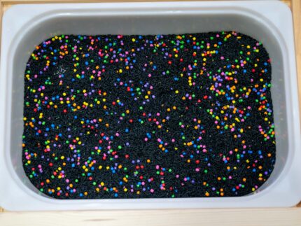Trick-or-Treat Sensory Bin with black rice, rainbow couscous, pumpkin containers, pretend candy, ghosts, pom-poms, and wooden scoop