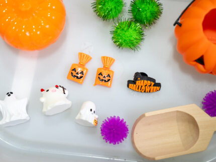 Trick-or-Treat Sensory Bin with black rice, rainbow couscous, pumpkin containers, pretend candy, ghosts, pom-poms, and wooden scoop