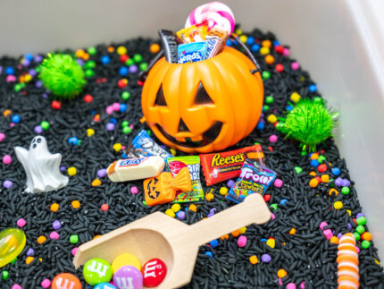 Trick-or-Treat Sensory Bin with black rice, rainbow couscous, pumpkin containers, pretend candy, ghosts, pom-poms, and wooden scoop