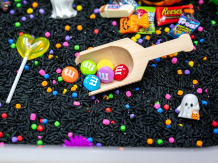 Trick-or-Treat Sensory Bin with black rice, rainbow couscous, pumpkin containers, pretend candy, ghosts, pom-poms, and wooden scoop