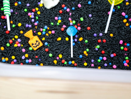 Trick-or-Treat Sensory Bin with black rice, rainbow couscous, pumpkin containers, pretend candy, ghosts, pom-poms, and wooden scoop