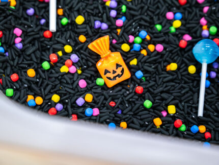 Trick-or-Treat Sensory Bin with black rice, rainbow couscous, pumpkin containers, pretend candy, ghosts, pom-poms, and wooden scoop