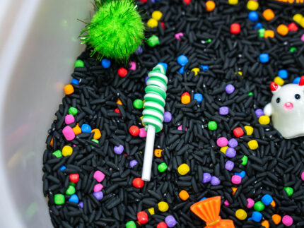 Trick-or-Treat Sensory Bin with black rice, rainbow couscous, pumpkin containers, pretend candy, ghosts, pom-poms, and wooden scoop