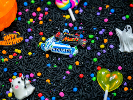 Trick-or-Treat Sensory Bin with black rice, rainbow couscous, pumpkin containers, pretend candy, ghosts, pom-poms, and wooden scoop