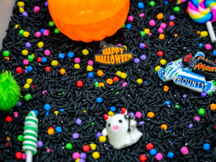 Trick-or-Treat Sensory Bin with black rice, rainbow couscous, pumpkin containers, pretend candy, ghosts, pom-poms, and wooden scoop