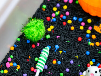 Trick-or-Treat Sensory Bin with black rice, rainbow couscous, pumpkin containers, pretend candy, ghosts, pom-poms, and wooden scoop